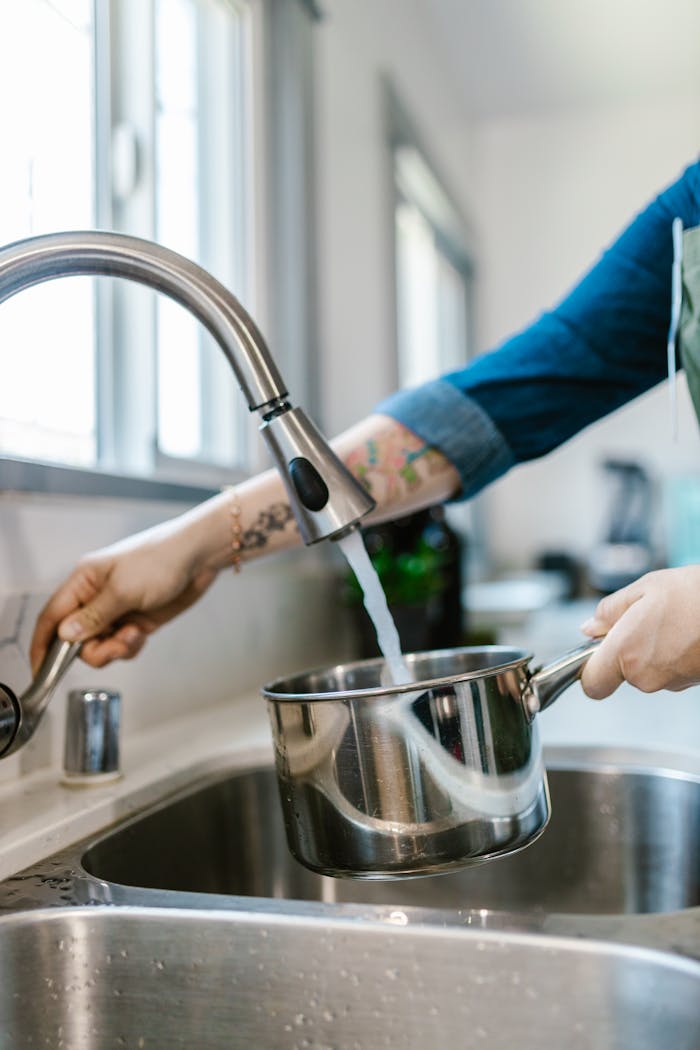 Hand filling a stainless steel pot with tap water in a modern kitchen.